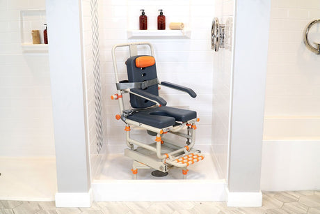 A specialized Showerbuddy chair designed for petite individuals, positioned inside a shower cubicle with white tiling and blue accents, featuring adjustable orange safety grips and footrests.