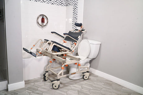 Showerbuddy bathroom transfer system featuring a tilt-in-space chair next to a standard toilet, set against a backdrop of white tiles and herringbone floor, highlighting accessible bathroom design.