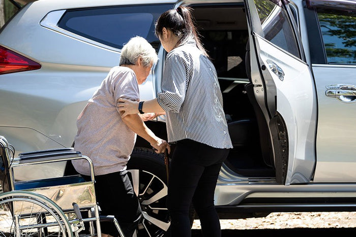 Image showing an elderly person being assisted into a car with a wheelchair in the background, titled.