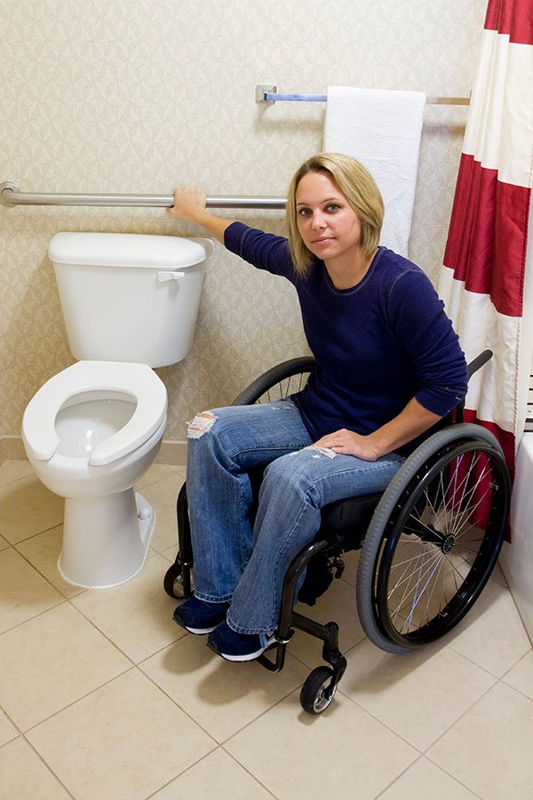 Image of a woman in a wheelchair in a bathroom, reaching for a grab bar, highlighting accessibility and mobility assistance in bathroom settings.