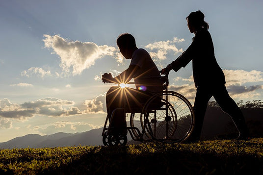 Silhouette of a person in a wheelchair and a caregiver outdoors at sunset, with the sun's rays creating a flare effect through the wheel spokes, symbolizing care and mobility support.