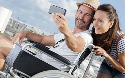 A couple taking a selfie, with one person seated in a wheelchair, symbolizing the ease of travel and holiday opportunities accessible with Showerbuddy's travel-friendly chairs.